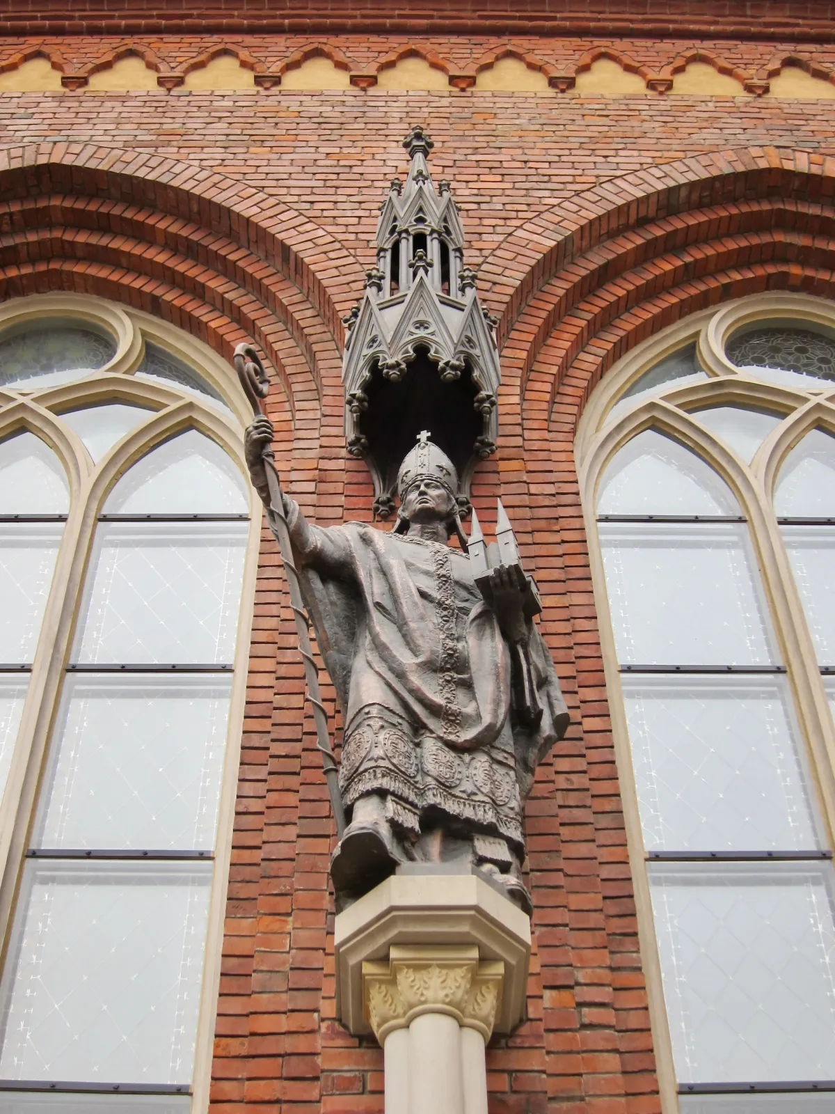 A bronze statue of a bishop holding a staff, set against a red brick church wall.