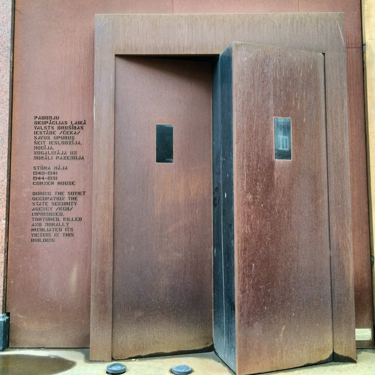 A heavy steel door on a brick wall, beside a memorial inscription in Latvian and English.