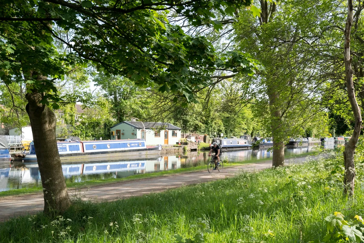 A cyclist on a canal towpath under trees, with narrowboats moored alongside.