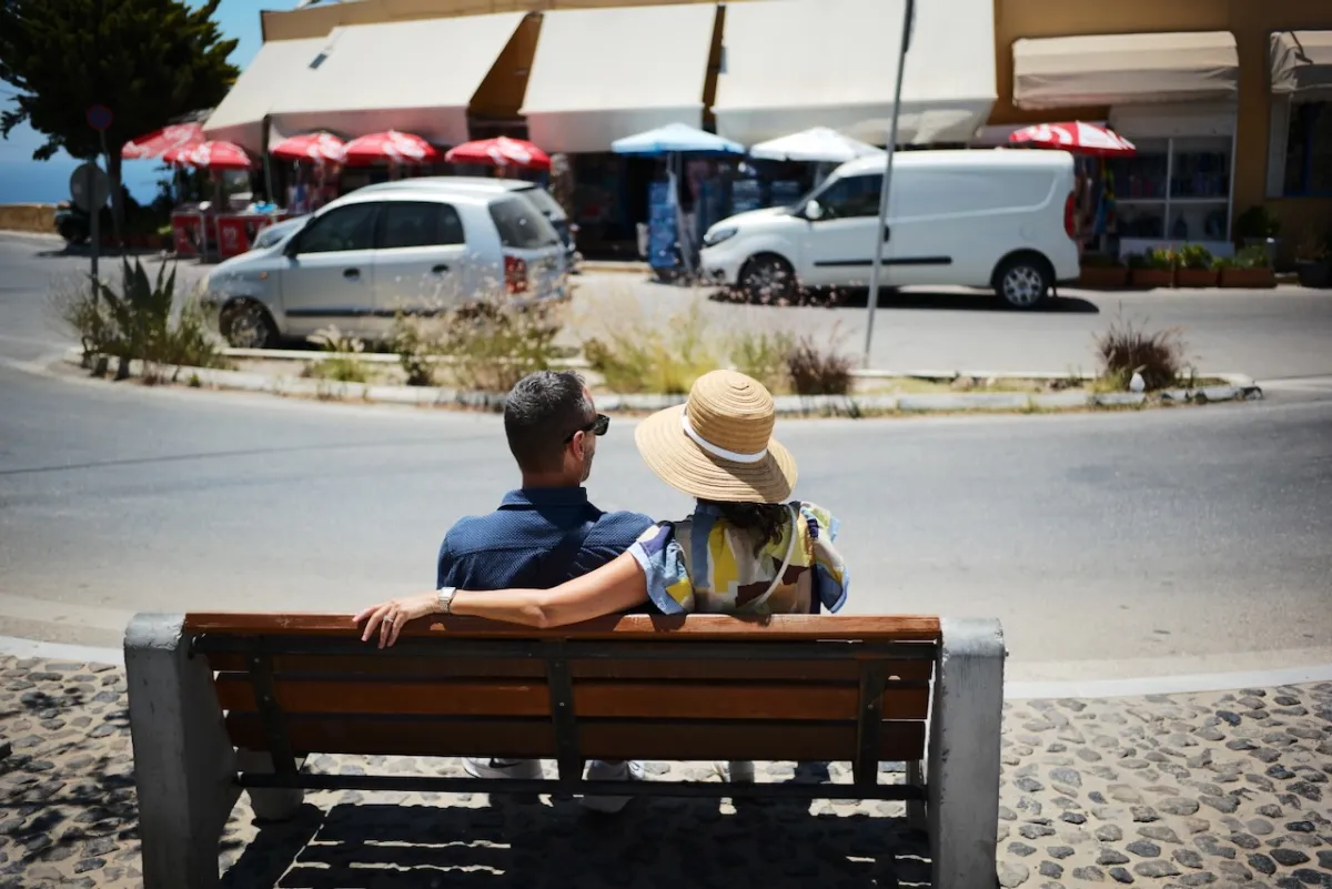 A couple sitting on a bench with their backs to the camera, looking across a street.