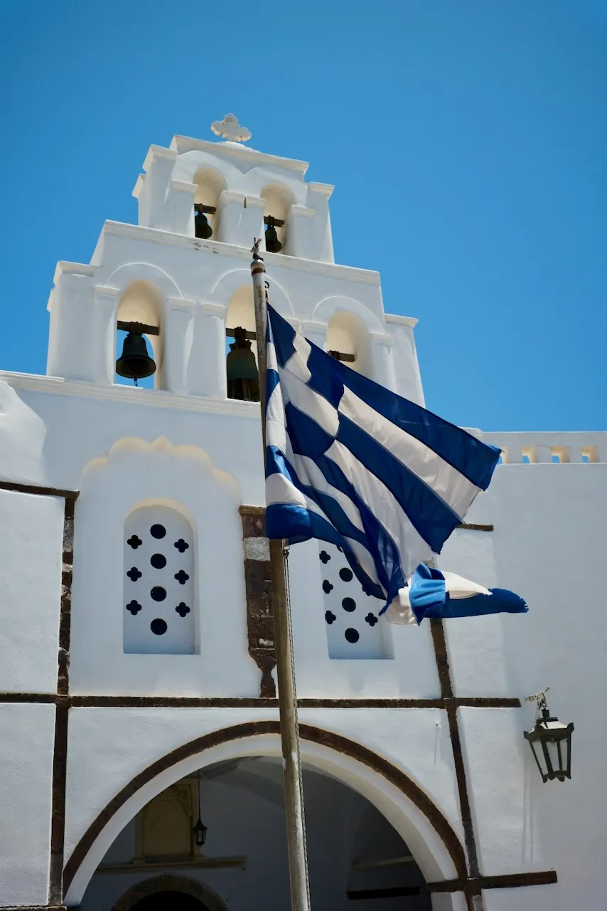 A Greek flag flying in front of a white church bell tower.