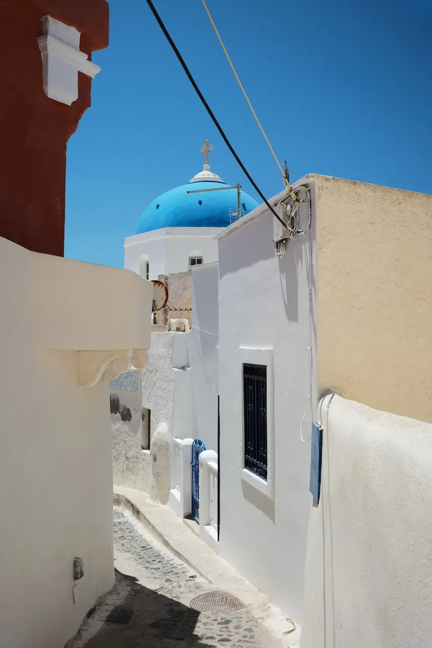 A narrow whitewashed alley leading towards a blue-domed church.