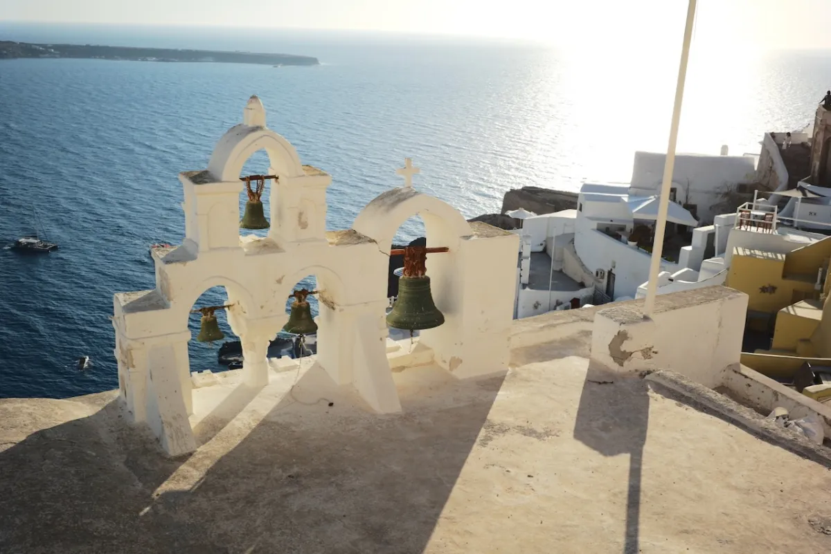 A whitewashed bell arch on a terrace above the sea, with the sun low over the water.