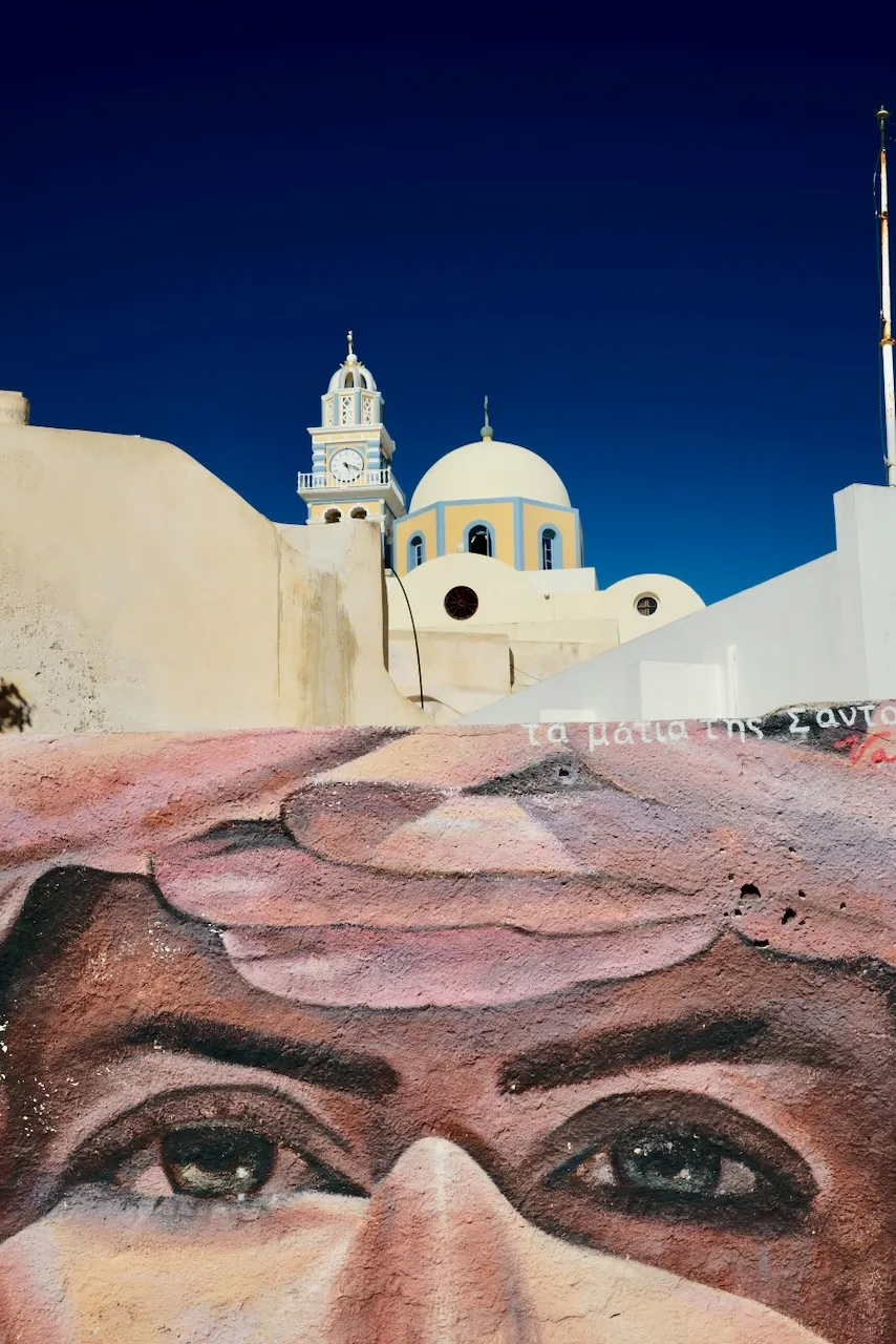 A large painted face on a wall, with a clock tower and domed church behind.