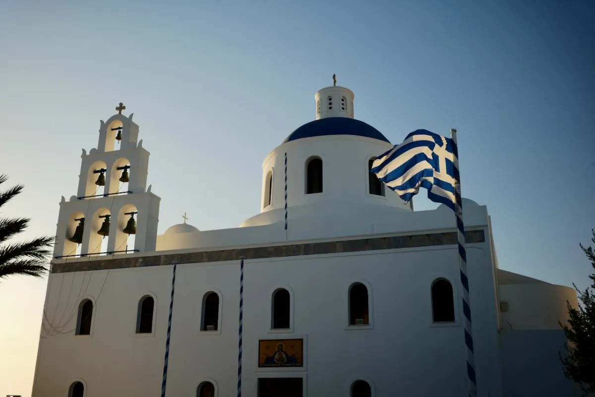 A white church with a blue dome and an open bellcote, with a Greek flag flying beside it.