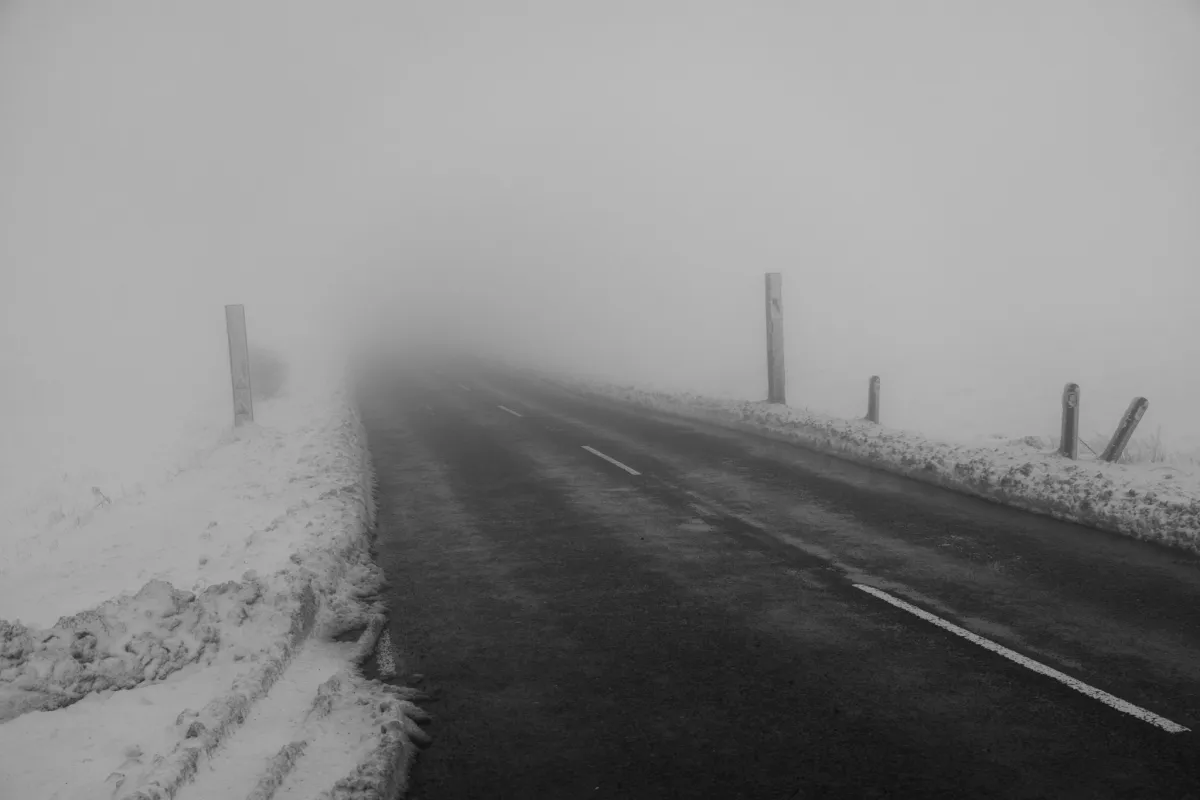 A road disappearing into fog, with snow piled along both verges. Black and white.