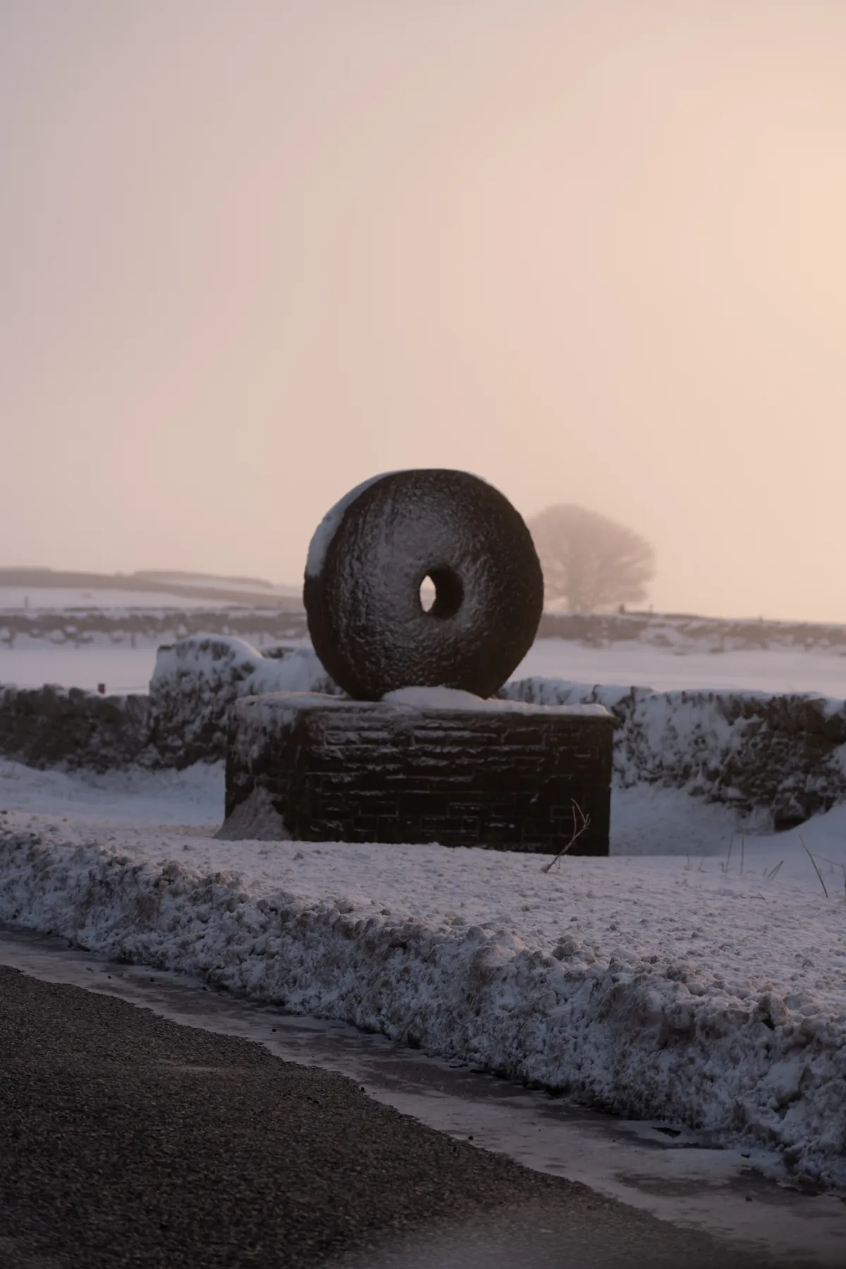 A large millstone mounted on a plinth in the snow, with mist behind.