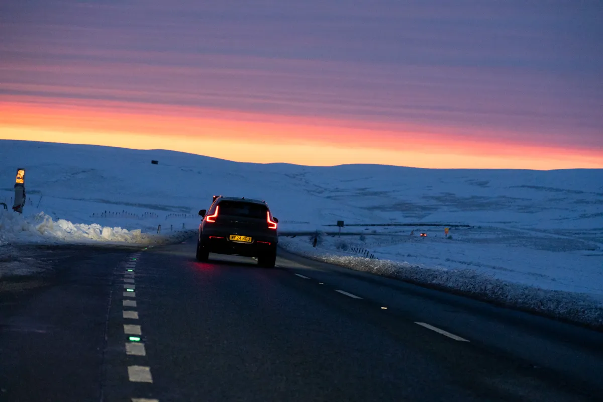 A car on a snow-lined road at sunset, with an orange sky ahead.