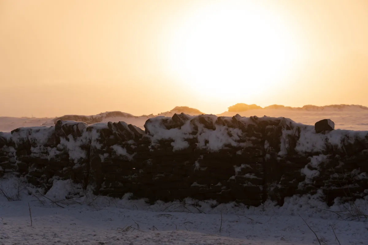 The sun low behind a snow-covered dry stone wall.