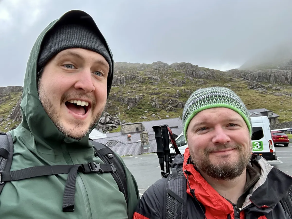 Two men in hats and waterproofs in a car park, with a rocky hillside behind them.
