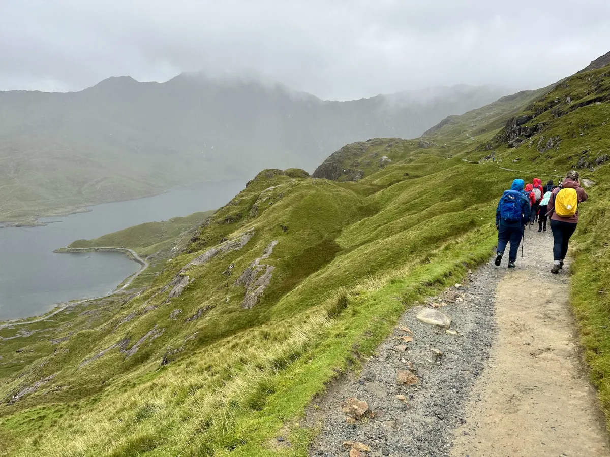 Walkers on a stony path along a steep grassy slope, with a lake far below.