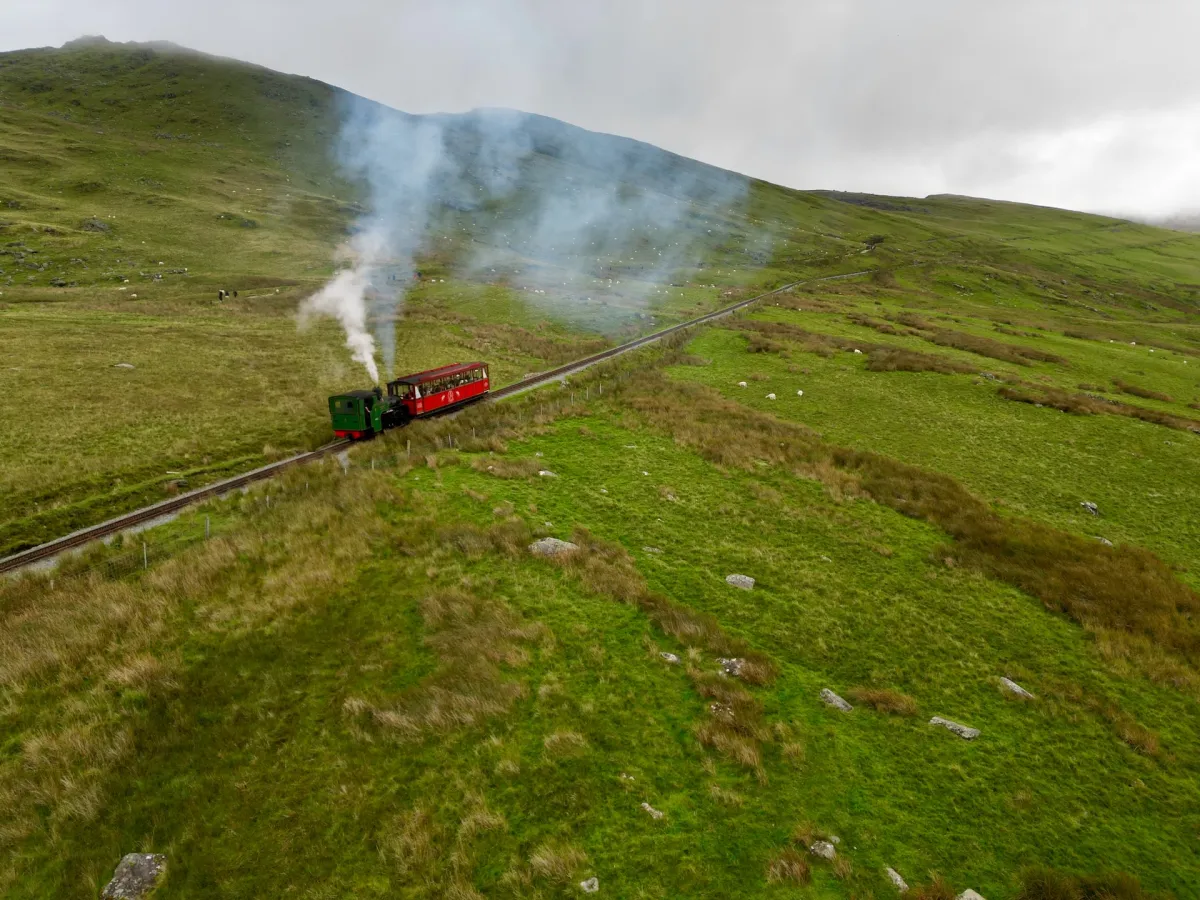 A steam locomotive pulling a single red carriage up a mountain railway across open hillside.
