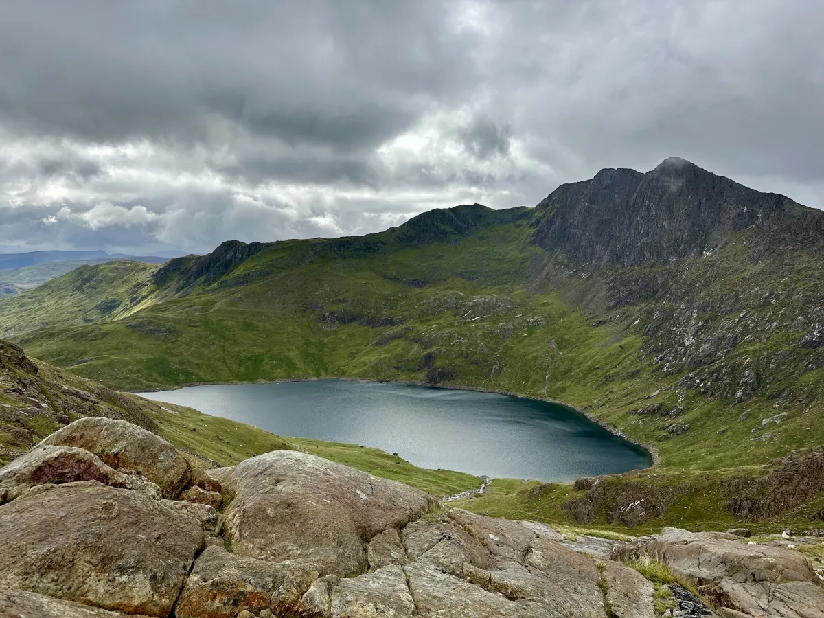 A dark mountain lake in a bowl of green and rocky slopes under heavy cloud.