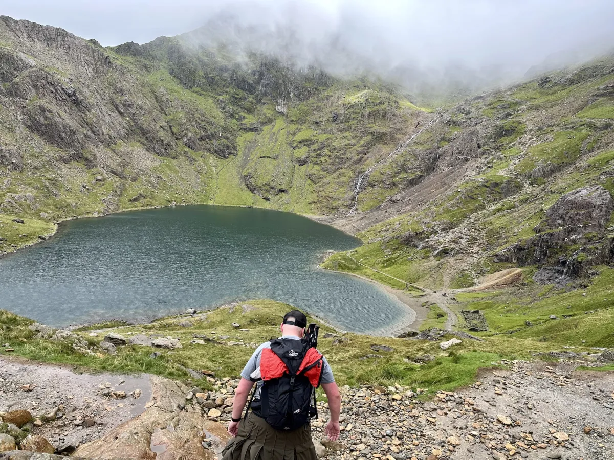 A walker with a red rucksack looking down over a mountain lake ringed by crags.