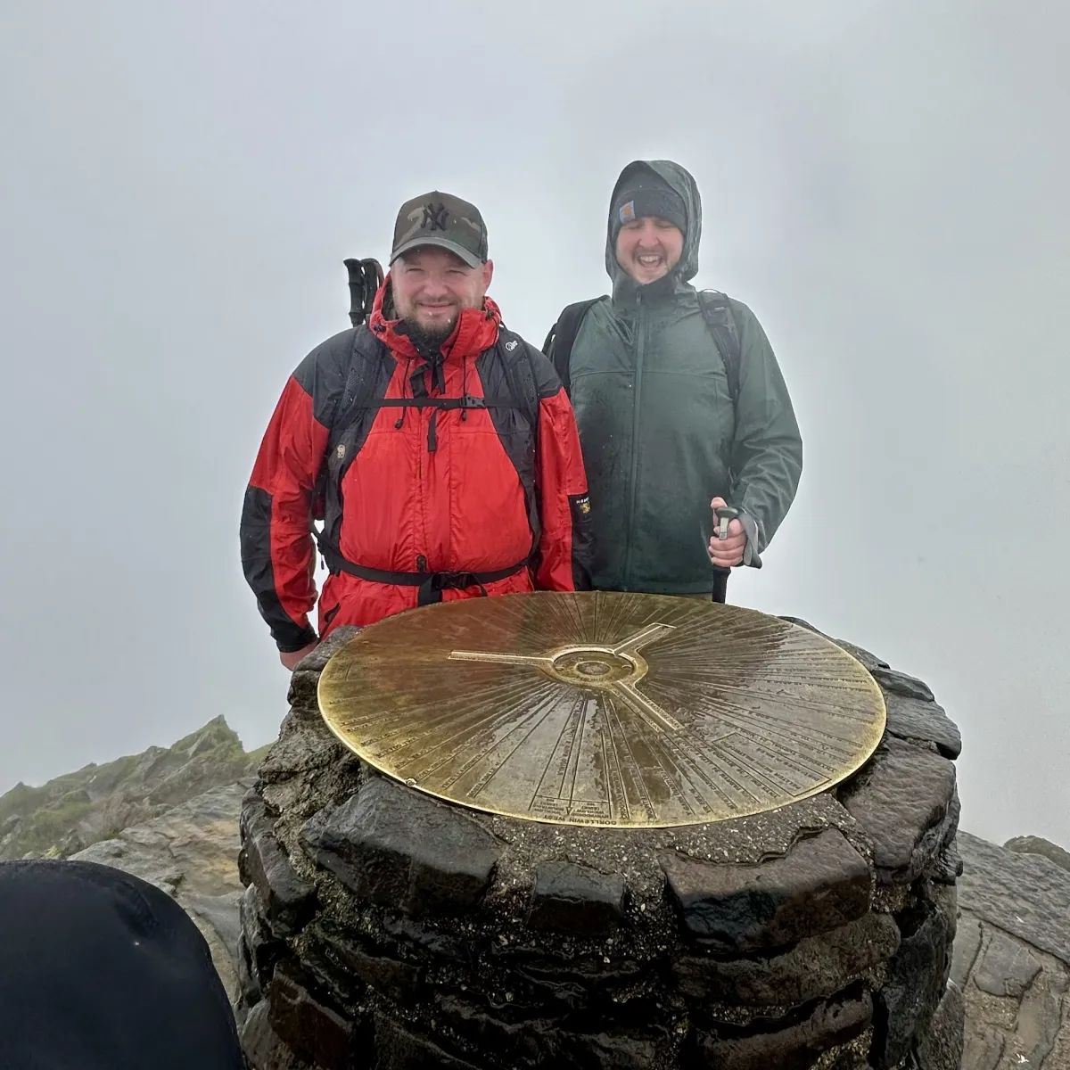 Two walkers in waterproofs standing behind the brass summit trig marker in mist.