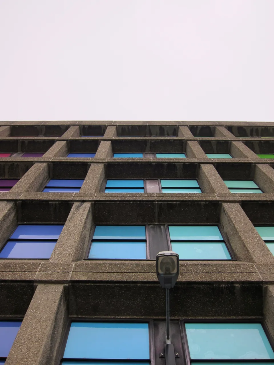 Looking up a concrete facade of blue and purple panels against a white sky.