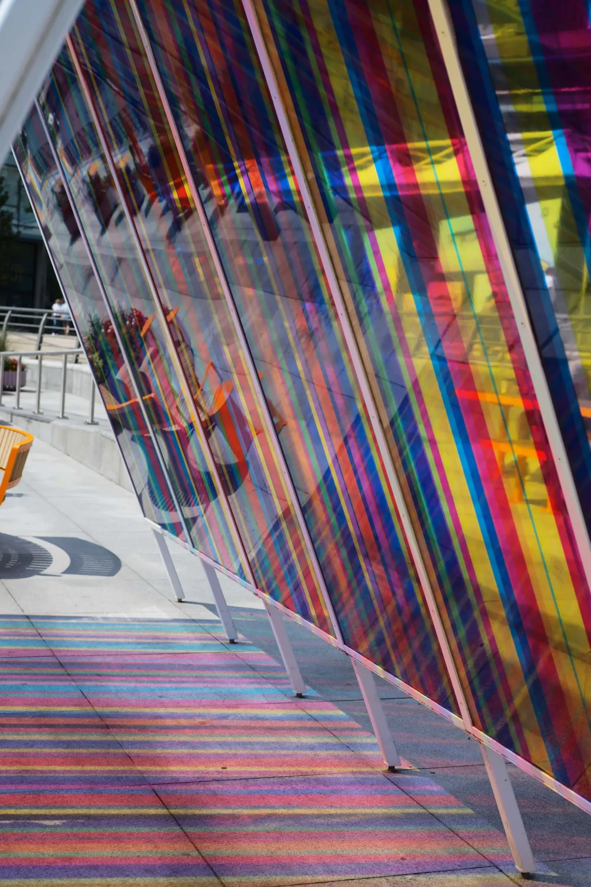 A walkway beside panels of coloured film casting striped shadows on the ground.