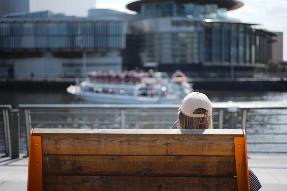 A person on a bench with their back to the camera, looking across water at a modern building.