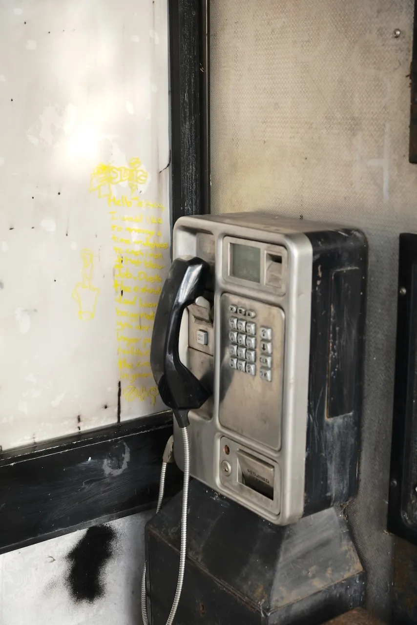A grimy payphone in a call box, with yellow marker scrawled on the panel behind.