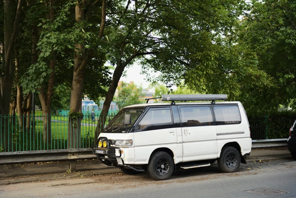 A white Mitsubishi camper van parked on a road beside railings and trees.