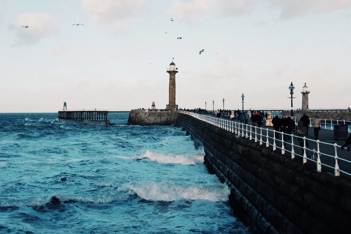 Waves breaking against a stone pier leading to a lighthouse, with people walking along it.