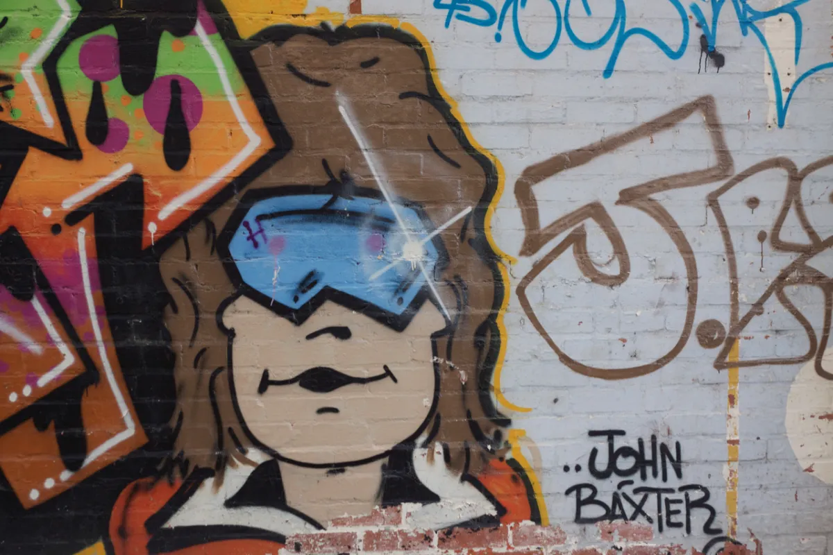 A painted face wearing blue ski goggles on a brick wall, beside other tags.