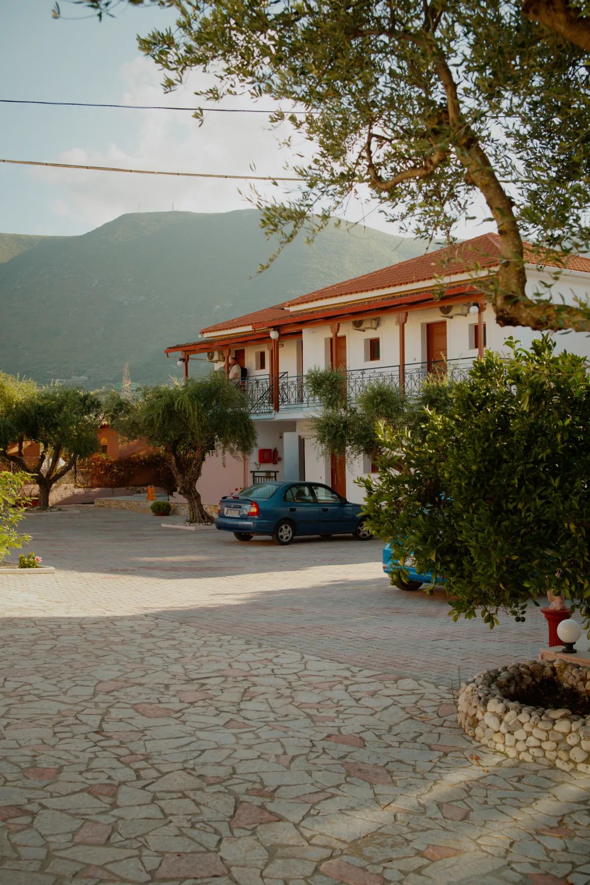 A white apartment building with balconies behind a paved forecourt, hills rising behind.