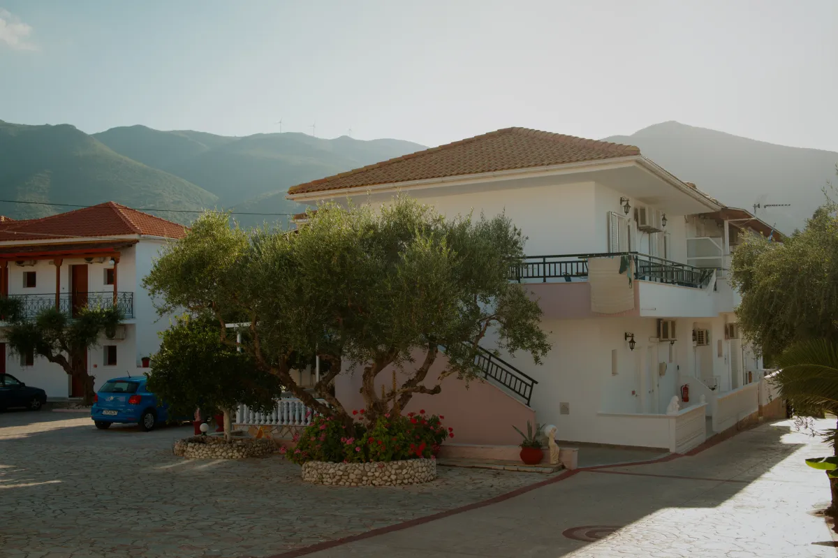 A white apartment building with balconies, olive trees in the forecourt and mountains behind.
