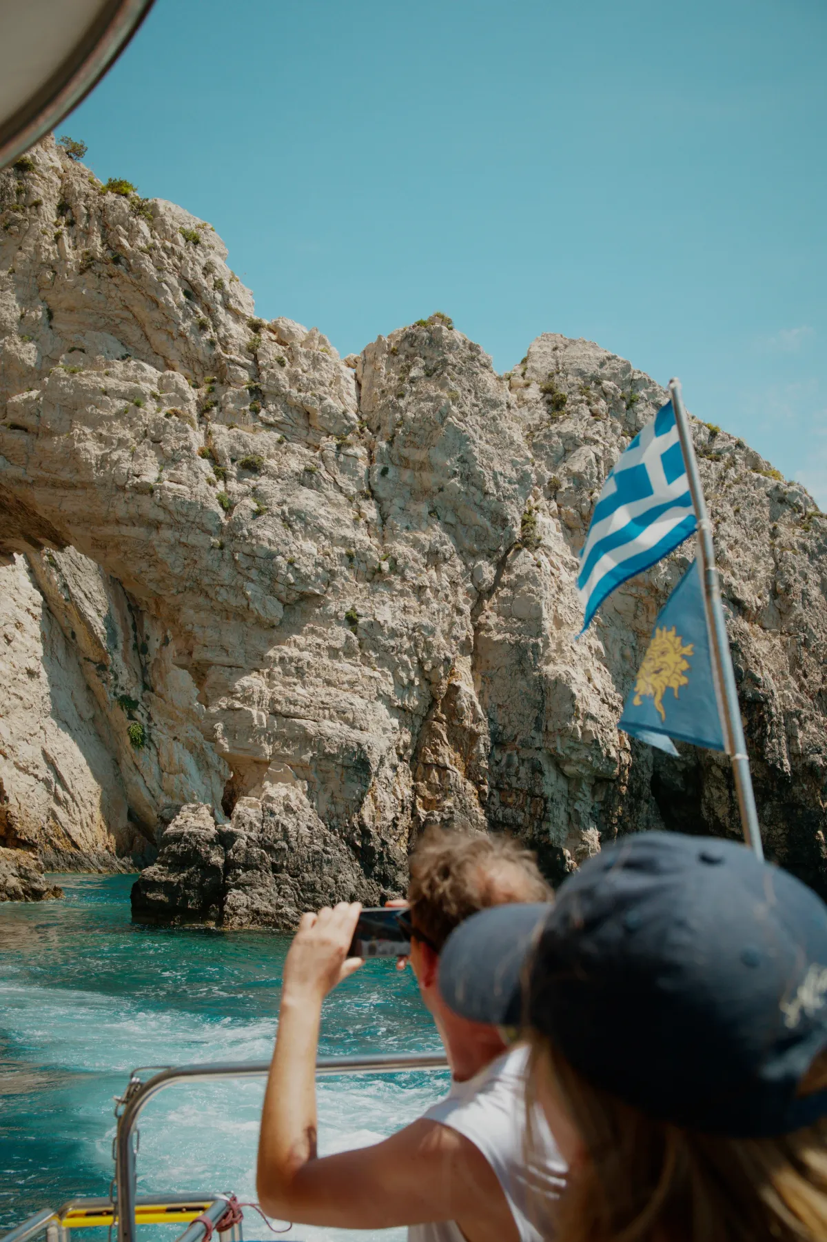 A passenger photographing the cliffs from a boat, with a Greek flag flying.