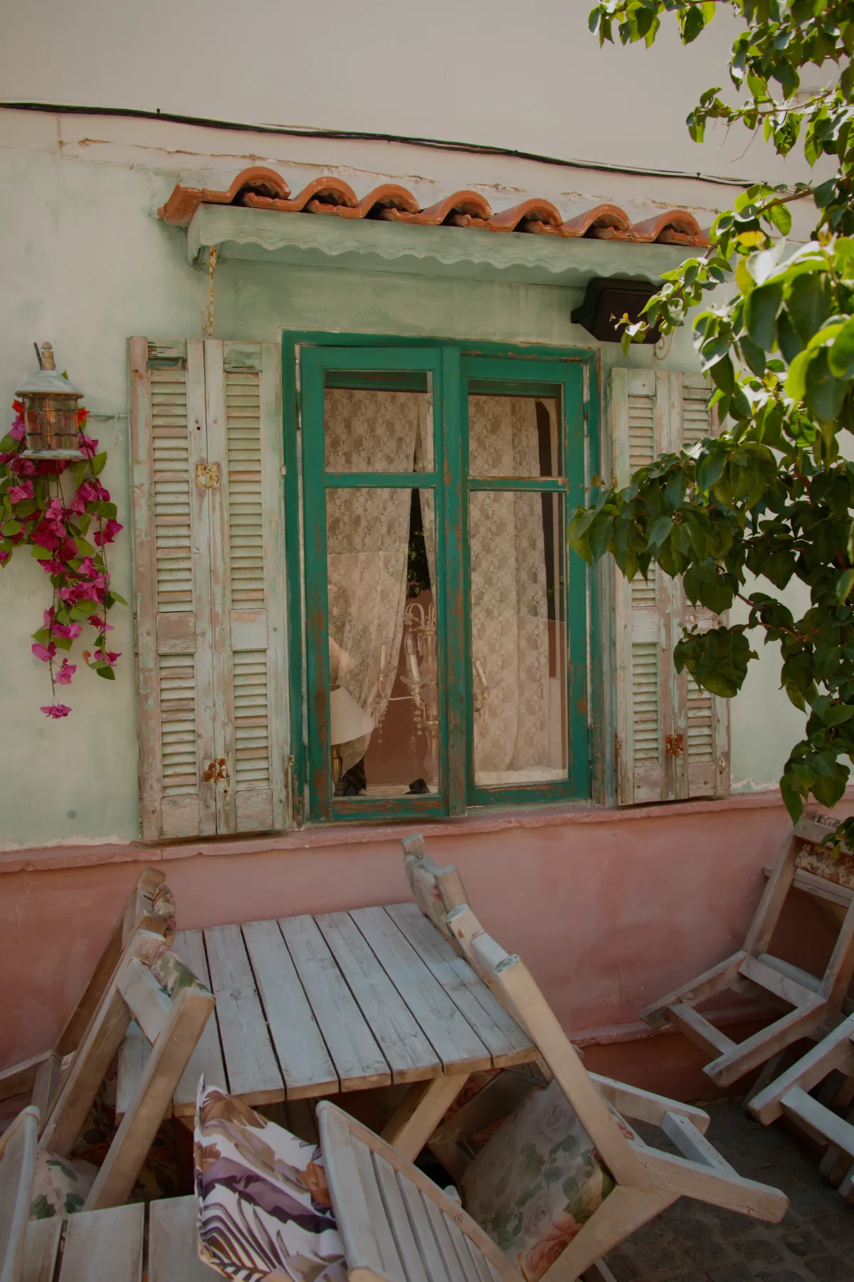 A green-framed window with old shutters, and a wooden table and chairs on the terrace.