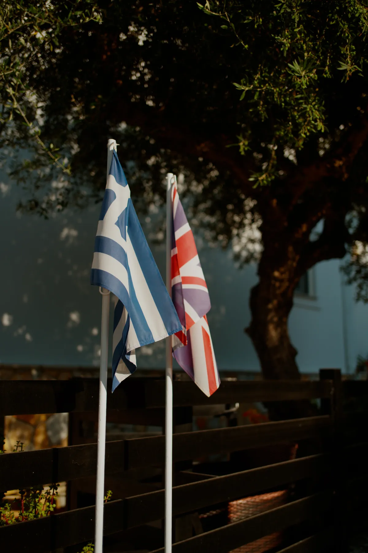 A Greek flag and a Union flag on poles beside a wooden fence.