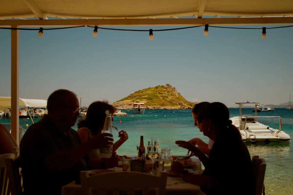 People eating at a beachside taverna under an awning, with an islet beyond.