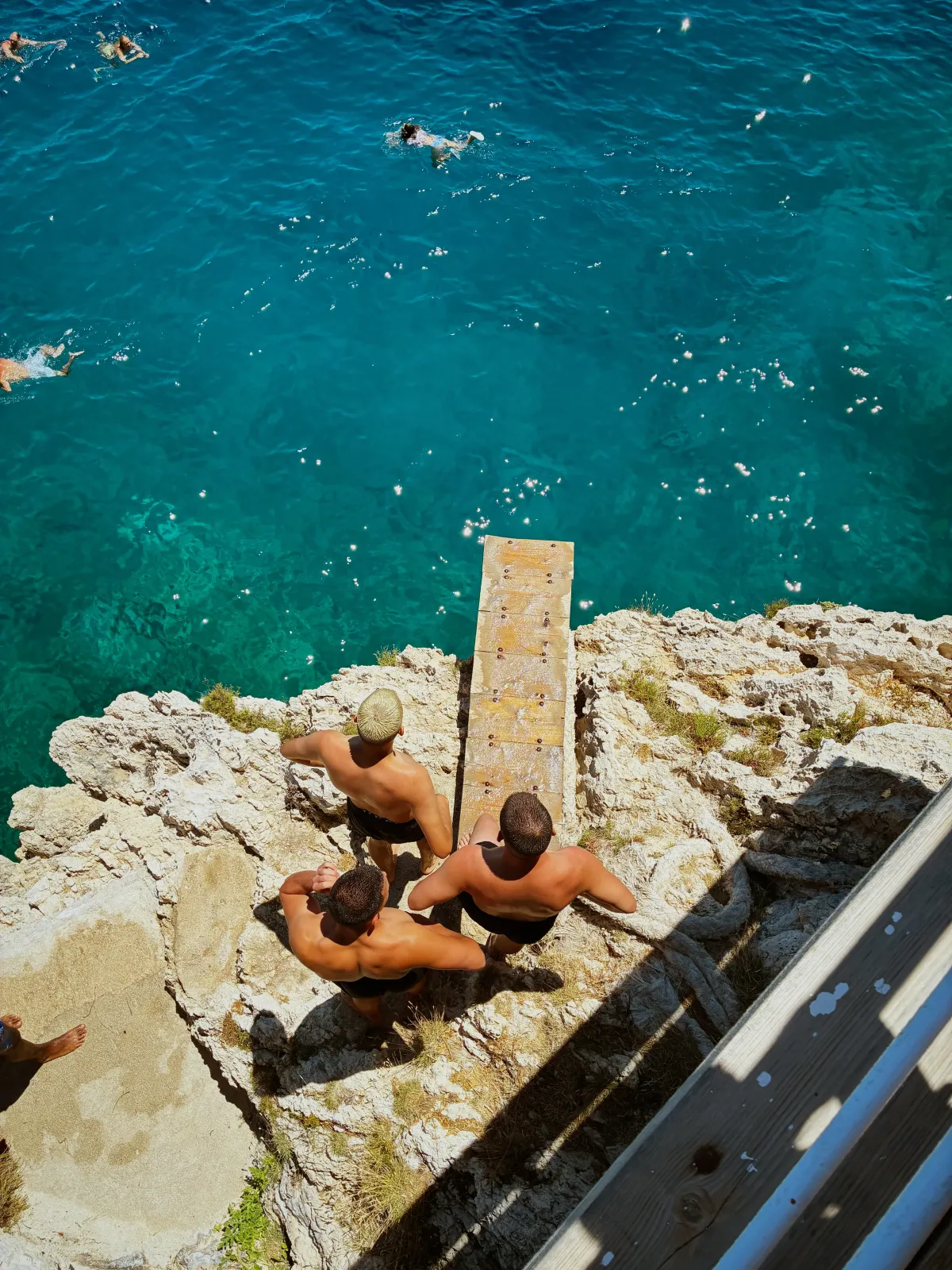 Three people sitting on rocks beside a diving board above clear water.