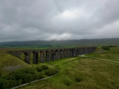 Aerial view of the Ribblehead Viaduct curving across green moorland, its stone arches stretching towards hills shrouded in low grey cloud