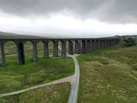 Drone shot looking along the arches of the Ribblehead Viaduct as a narrow lane passes beneath them, under a heavy overcast sky